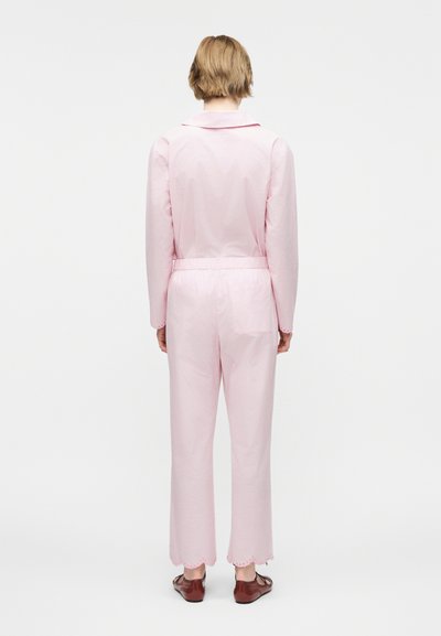 Άτομο με κοντά ξανθά μαλλιά, ντυμένο με ανοιχτό ροζ μακρυμάνικη ολόσωμη φόρμα και καφέ παπούτσια, στέκεται με την πλάτη προς το μέρος μας πάνω σε λευκό φόντο.