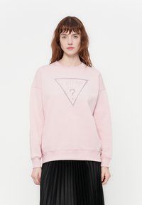 Mujer con cabello rojo vistiendo un sudadera rosa claro de Guess y una falda negra plisada contra un fondo blanco liso.