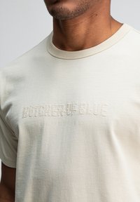 Close-up van een man die een beige t-shirt draagt met geborduurd de tekst "BUTCHER OF BLUE" en in lichte belettering "QUALITY CLOTHING CO" op de borst.