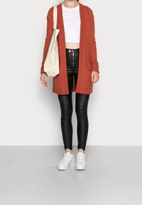 Persona che indossa un cardigan lungo color ruggine, top corto bianco, pantaloni in pelle nera, sneakers bianche e porta una borsa a tracolla beige sulla spalla sinistra.