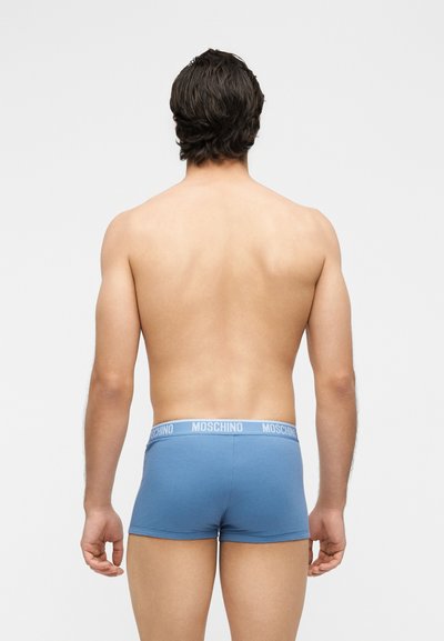 Άνδρας με κοντά σκούρα μαλλιά, ξυπόλυτος, χωρίς πουκάμισο, φοράει μπλε μποξεράκια Moschino, με την πλάτη γυρισμένη προς ένα απλό ανοιχτό φόντο.