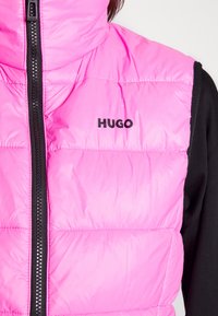 Helle pink gesteppte Weste mit schwarzem Reißverschluss und "HUGO"-Logo auf der Brust, getragen über einem schwarzen Langarmshirt.