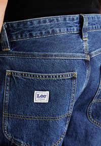 Blue denim jeans with a distinct patch featuring "Lee WORKWEAR," yellow stitching, and a classic five-pocket design.
