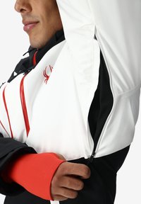 Giacca bianca e nera con dettagli rossi, dotata di tasca con cerniera e fodera in rete. Il tessuto ha una consistenza liscia.