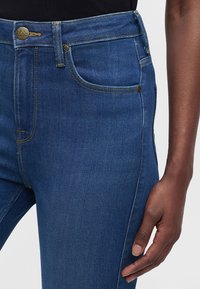 Gros plan sur un jean en denim bleu avec un bouton en laiton et des coutures, porté par une personne à la peau foncée portant un haut noir.