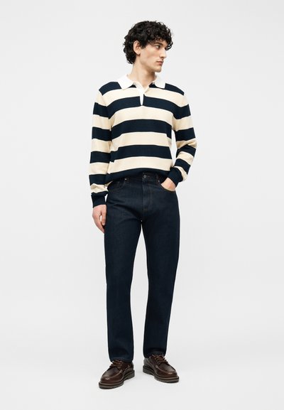 Άνδρας με σγουρά μαλλιά, φοράει μακρυμάνικο μπλουζάκι πόλο με κρεμ και ναυτικές ρίγες, σκούρα τζιν παντελόνια και καφέ δερμάτινα παπούτσια, στέκεται με το ένα χέρι στην τσέπη.