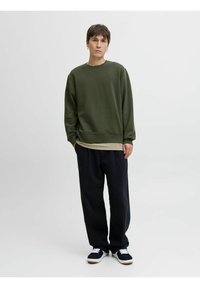 Olijfgroen sweatshirt met lange mouwen, ronde hals en geribbelde zoom, gelaagd over een lichtbeige shirt. Gecombineerd met losse zwarte broek en sneakers.
