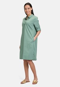 Kurzärmliges, kragenloses Kleid aus hellgrünem Stoff mit seitlichen Taschen, knielangem Design und aufgerollten Ärmeln. Getragen mit goldenen Riemchensandalen.