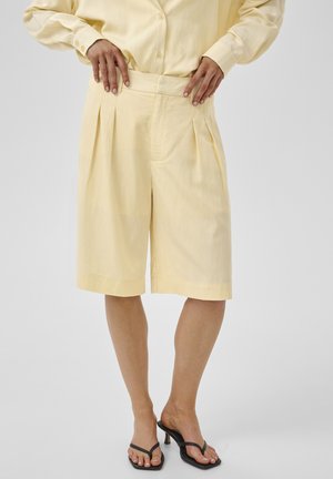 Persoon draagt losse, knie-lange lichtgele shorts met plooien, gecombineerd met een bijpassend shirt en zwarte sandalen met hak.