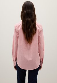 Donna con lunghi capelli castani che indossa una camicia a maniche lunghe rossa e bianca a righe verticali e pantaloni scuri, vista da dietro.