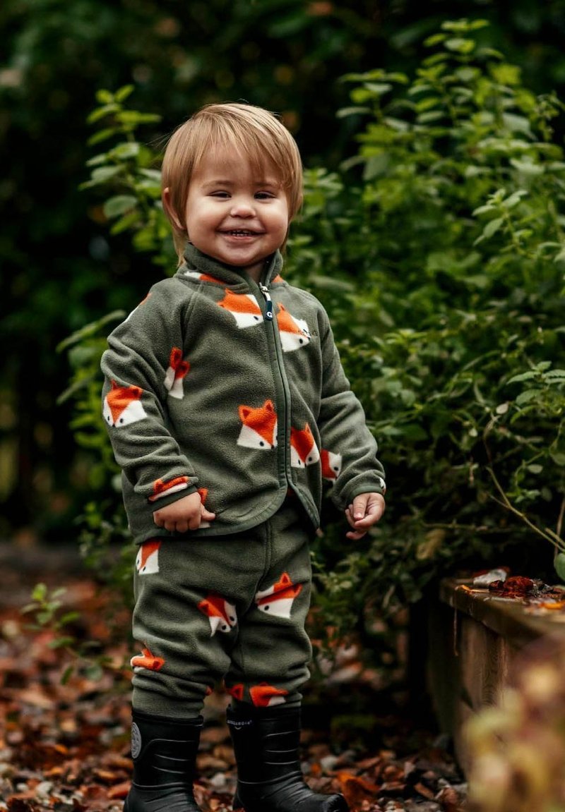 Grön fleece-outfit med orange rävmönster, zip-up jacka och matchande byxor. Svarta gummistövlar fulländar looken.