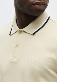 Homme portant un polo beige avec un col texturé et une bordure noire et blanche, logo "BOSS" brodé sur la poitrine.