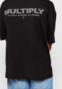 Černé oversize tričko s velkým šedým nápisem "MULTIPLY APPAREL CLUB 2023" na zadní straně, vyrobené z měkké bavlny.