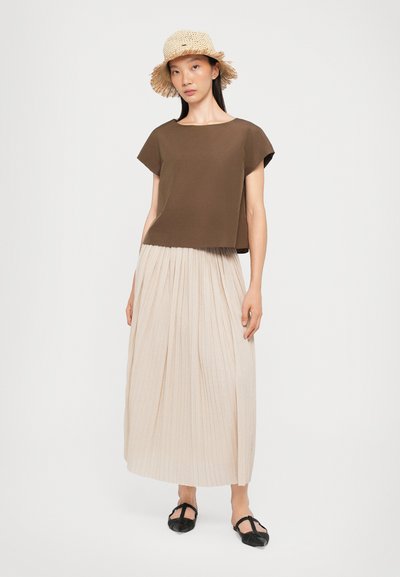 Haut marron à manches courtes avec un col rond, associé à une jupe maxi plissée beige. Des chaussures plates noires complètent la tenue.