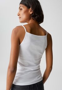 Woman with short dark hair wearing a white sleeveless top and dark jeans, looking to her left against a plain background.