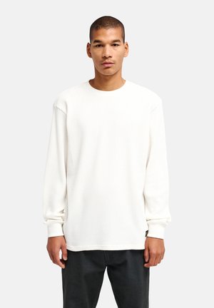 Jeune homme aux cheveux courts portant une chemise blanche texturée à manches longues et un pantalon noir, debout devant un fond clair uni.
