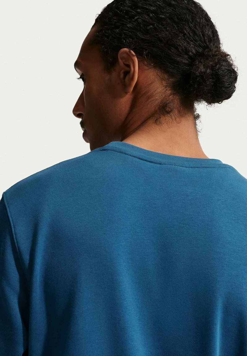 Felpa blu con scollo rotondo, tessuto morbido e dettaglio cucitura sulla schiena, indossata da una persona con capelli ricci raccolti dietro.