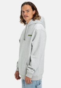 Sudadera con capucha gris, bolsillo canguro, capucha con cordón y acento en verde del logo en el pecho. Tela suave y corte relajado.