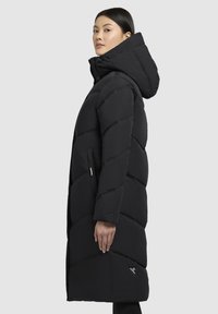 Geacă puf neagră cu glugă, având un design matlasat, buzunare laterale și un accent cu logo pe tiv. Material texturat, izolat.