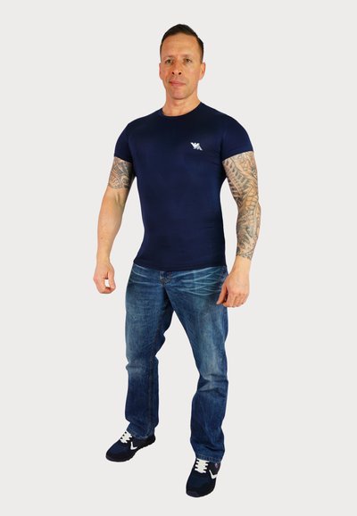 Marineblaue, taillierte T-Shirt mit kurzen Ärmeln, das ein kleines Logo trägt. Kombiniert mit hellblauen Jeans und schwarzen Sportschuhen.