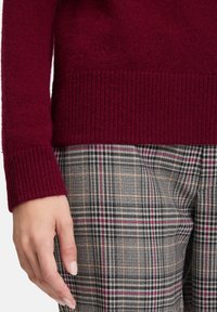 Hand med manikyrerade naglar bredvid rutig grå, svart och rosa byxor samt ribbad mudd av vinröd tröja.