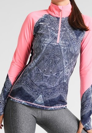 Kobieta w sportowej bluzce z długim rękawem, z różowymi rękawami i niebieskim wzorzystym przodem, w połączeniu z szarymi legginsami.