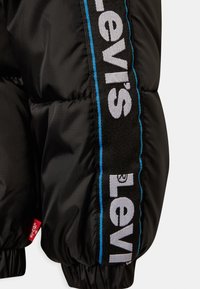 Giacca a vento nera con una prominente striscia nera e blu e logo "Levi's", tessuto testurizzato e polsini elasticizzati nella parte inferiore.