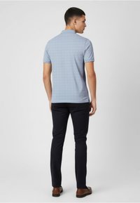 Camisa polo azul claro con un patrón sutil, mangas cortas y cuello, combinada con pantalones oscuros y zapatos marrones, vista desde atrás.