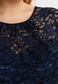 Haut en dentelle bleu marine avec un col rond, des détails froncés aux épaules et un motif floral complexe sur l'ensemble.