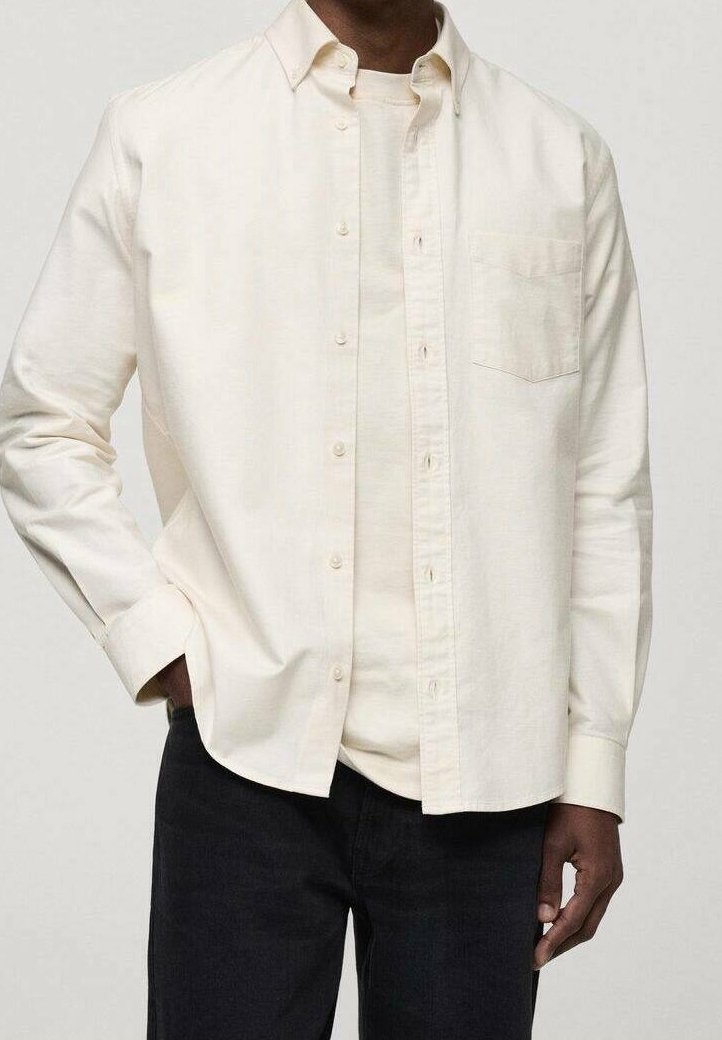 Homme portant une chemise à manches longues écrue déboutonnée sur un t-shirt écru assorti et un pantalon noir, main dans la poche.