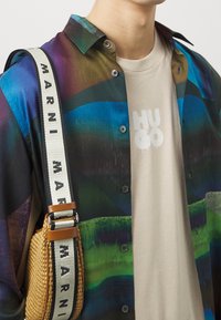 Multicolored button-up shirt featuring abstract patterns in green, blue, and purple, layered over a beige t-shirt with "HUGO" printed in white. A woven bag with brown accents and a strap labeled "MARNI" completes the look.