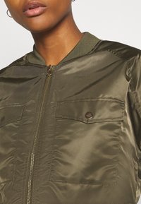 Veste bomber vert olive en tissu lisse, avec une fermeture éclair à l'avant et deux poches poitrine boutonnées. Col et poignets côtelés.