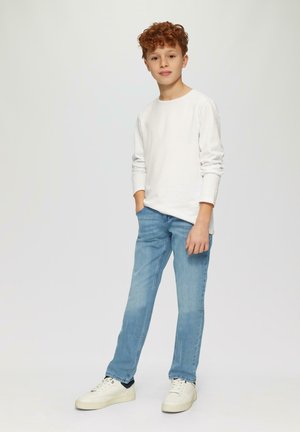 Weißes Langarmshirt, hellblaue Jeans und weiße Sneakers. Der Junge steht mit den Händen in den Taschen und zeigt einen lässigen Stil und eine sportliche Figur.