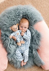 Light blue onesie with navy stars, plush teddy bear, and fluffy gray blanket. Soft texture and rounded shape with pink accents in the background.