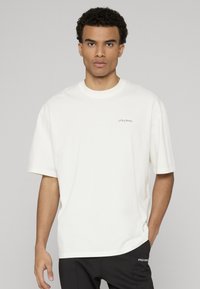 Valkoinen puuvillainen t-paita, jossa on pyöreä pääntie ja lyhyet hihat. Edessä pieni musta logo. Sileä pinta ja rento istuvuus.