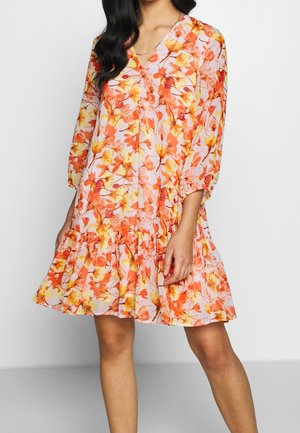 Mujer con un vestido hasta la rodilla, de manga larga, con estampado floral en tonos naranjas y amarillos y dobladillo con volantes, de pie frente a un fondo liso.