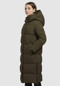 Long, puffer coat in dark olive green with a hood, featuring rectangular quilted sections and zippered side pockets on a smooth fabric.