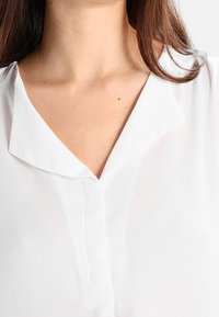 Blusa blanca con escote en V y cuello, hecha de una tela ligera y transparente, que presenta una tapeta delantera y una textura suave.