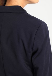 Námořnický blejzr s klopaným límcem, strukturovanými rameny a hladkým, texturovaným materiálem. Viděn ze zadní strany, ukazuje elegantní linie.