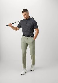 Άνδρας με ριγέ μαύρο και γκρι πόλο, ανοιχτό πράσινο παντελόνι, λευκά sneakers, κρατώντας ένα γκολφ κλαμπ στον ώμο, ενώ στέκεται μπροστά σε απλό φόντο.