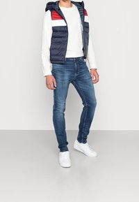 Chaleco acolchado azul marino con detalles en rojo y blanco sobre una camisa blanca de manga larga, combinado con jeans azules y zapatillas blancas. Silueta ajustada.