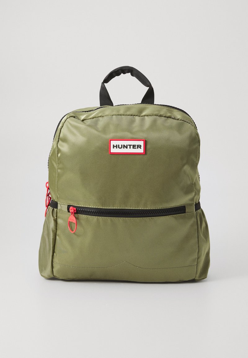 Olíva zöld nylon hátizsák fekete kiegészítőkkel, elől cipzáras zsebbel, piros cipzárhúzóval és egy kis logóval a cipzár felett.