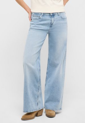 Osoba ubrana w jasnoniebieskie dżinsy o szerokich nogawkach i beżowe zamszowe kozaki na obcasie, stojąca z jedną ręką opartą na biodrze.