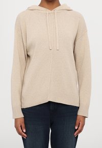 Sweat à capuche en maille beige avec poignets et ourlet côtelés, porté avec un jean bleu foncé, visible du cou jusqu'aux hanches sur fond blanc.