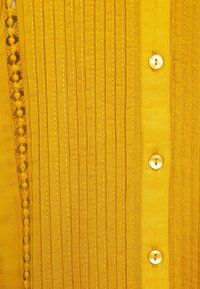 Hořčicově žlutá košile na knoflíky s vertikálním prošíváním a krajkovými detaily podél okraje. Obsahuje kulaté knoflíky a hladkou strukturu.