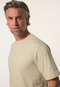 Helles beiges T-Shirt mit Rundhalsausschnitt aus weichem Stoff, mit kurzen Ärmeln und einem dezenten Logo auf dem linken Ärmel.