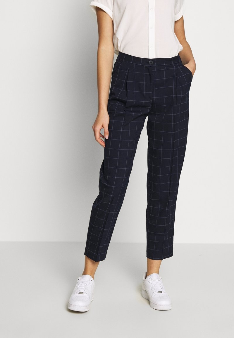 Femme portant un pantalon court à carreaux bleu marine, des baskets blanches et une chemise blanche à manches courtes boutonnée, les mains dans les poches.