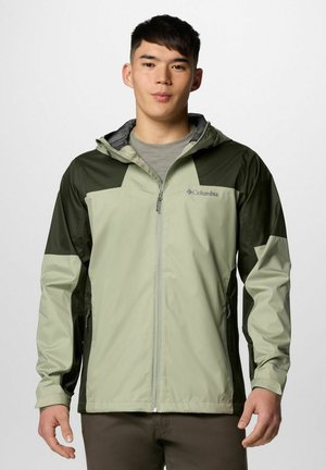 Junger Mann trägt eine hell- und dunkelgrüne Columbia Zip-up-Jacke mit Kapuze, steht vor einem schlichten weißen Hintergrund.