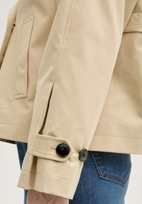 Beige leichte Jacke mit schwarzen Knöpfen, Schulterklappen und einer Brusttasche; teilweise an einer Person mit blauen Jeans gezeigt.