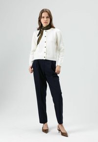Cardigan blanc en maille avec encolure ronde et fermeture à boutons, associé à un pantalon bleu marine sur mesure et une écharpe noire à motifs. Chaussures marron à talons.